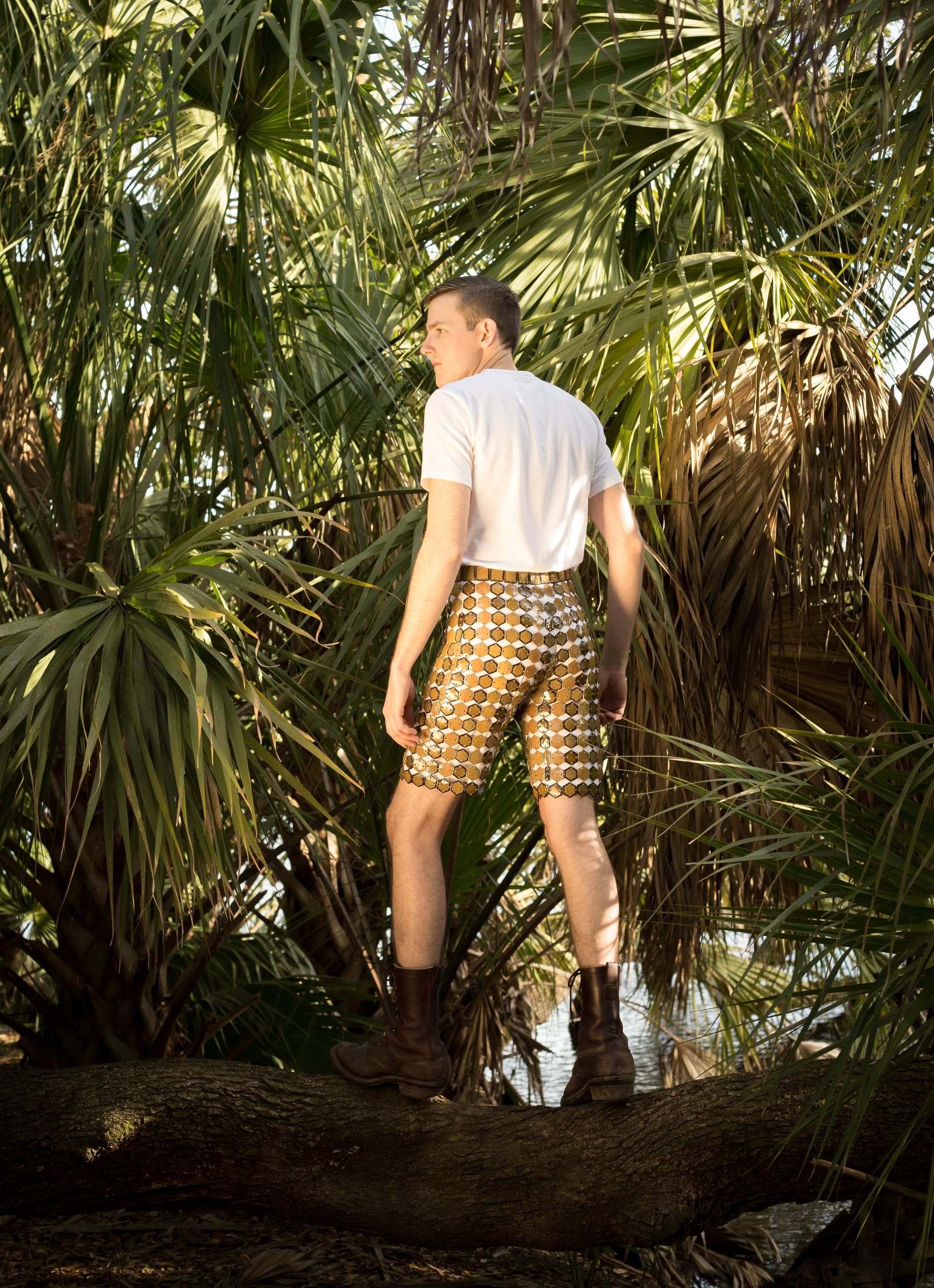 Shae Bishop wears gold and brown tile shorts with a white t-shirt and boots in a jungle-like setting