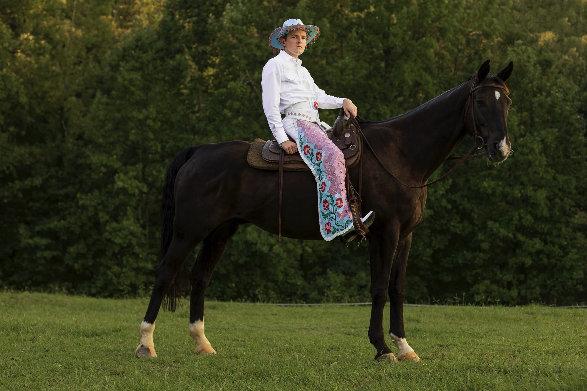 Shae Bishop wears a floral cowboy hat with red tassels on the edge of the brim, while seated on a horse. he also wears floral chaps made of tile