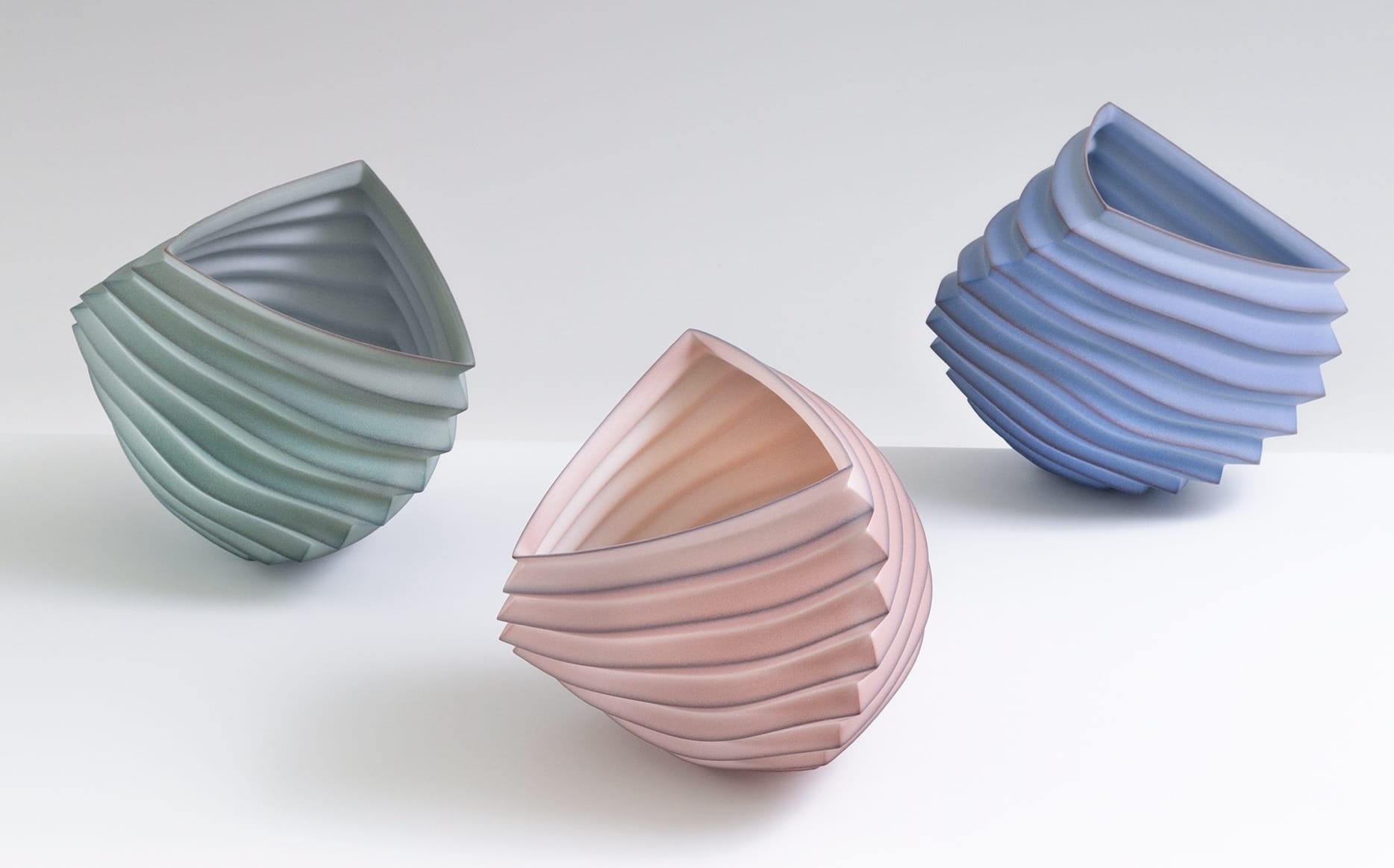 three abstract porcelain vessels loosely resembling intricately folded paper in pastel green, pink, and blue