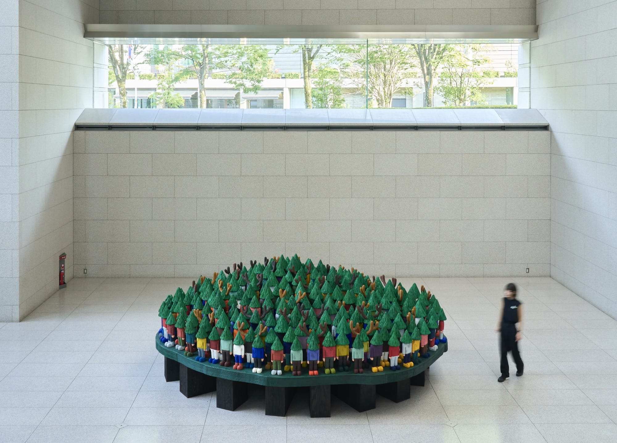 a sculptural installation in a contemporary museum space by Yuichi Hirako of an oversized table covered with cartoonish figures with evergreen trees for heads