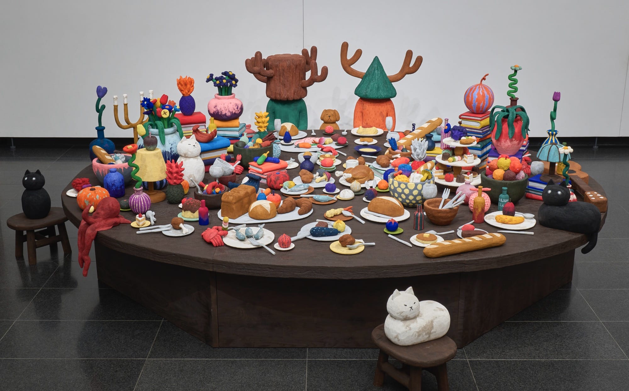 a sculptural installation by Yuichi Hirako of an oversized table covered with cartoonish domestic forms like vases, food, candlesticks, cats, and other objects