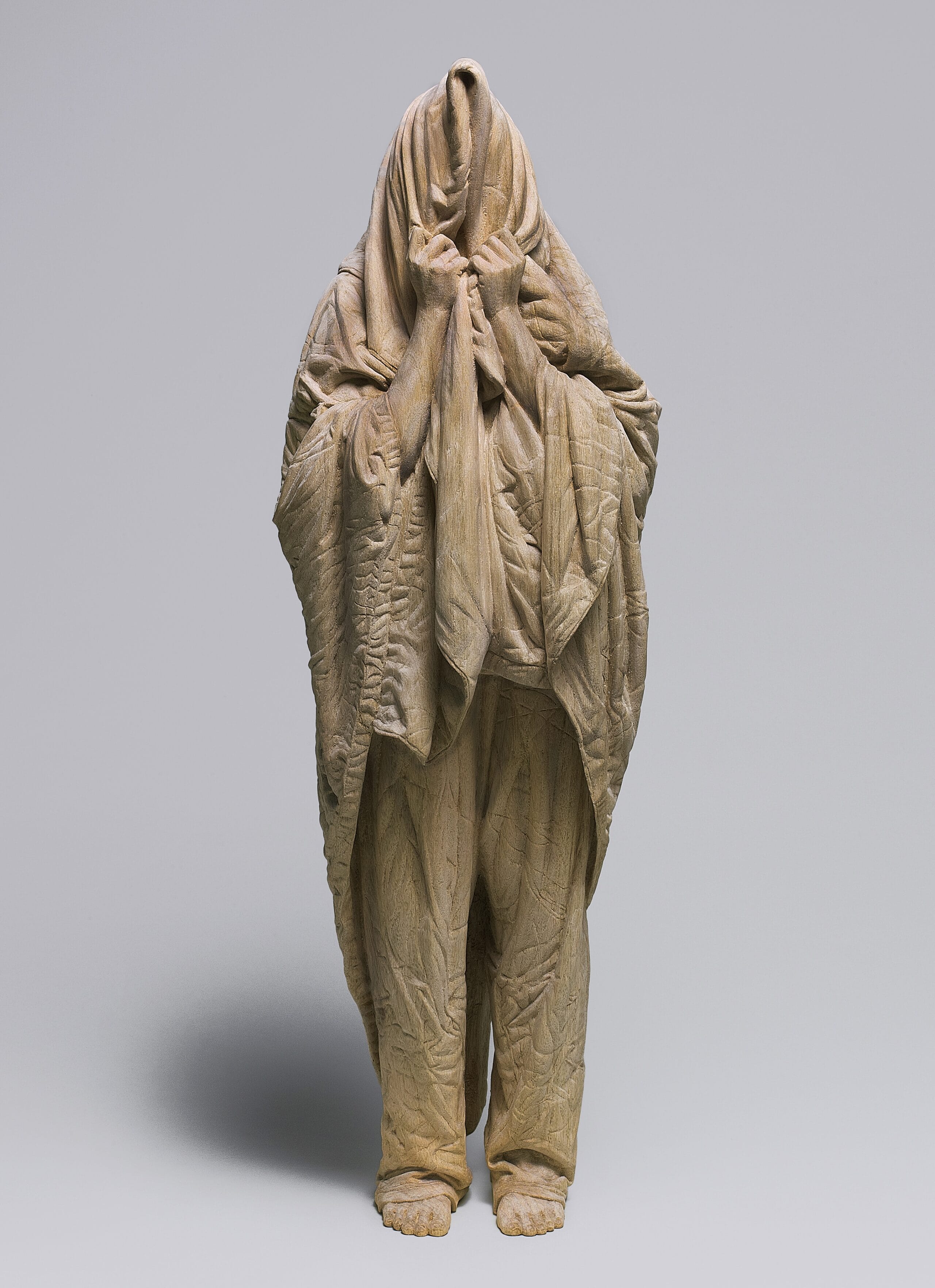 a carved wooden sculpture by Ikuo Inada of a figure standing with a blanket over their head as if very sleepy