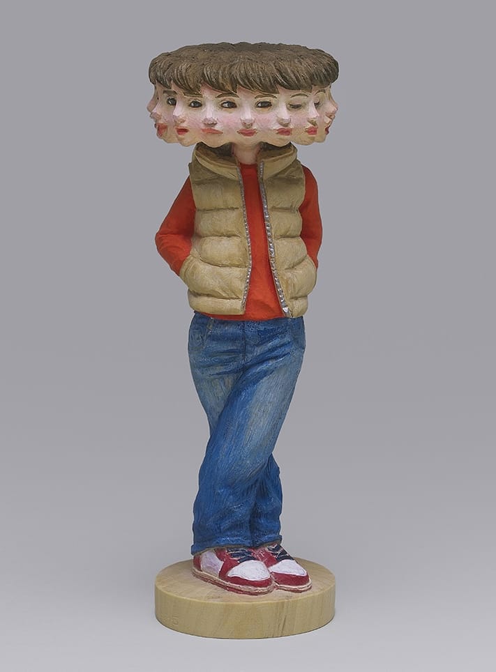 a wooden sculpture by Yoshitoshi Kanemaki of a woman wearing jeans and a red shirt and vest, with a head that appears to have numerous faces with different expressions