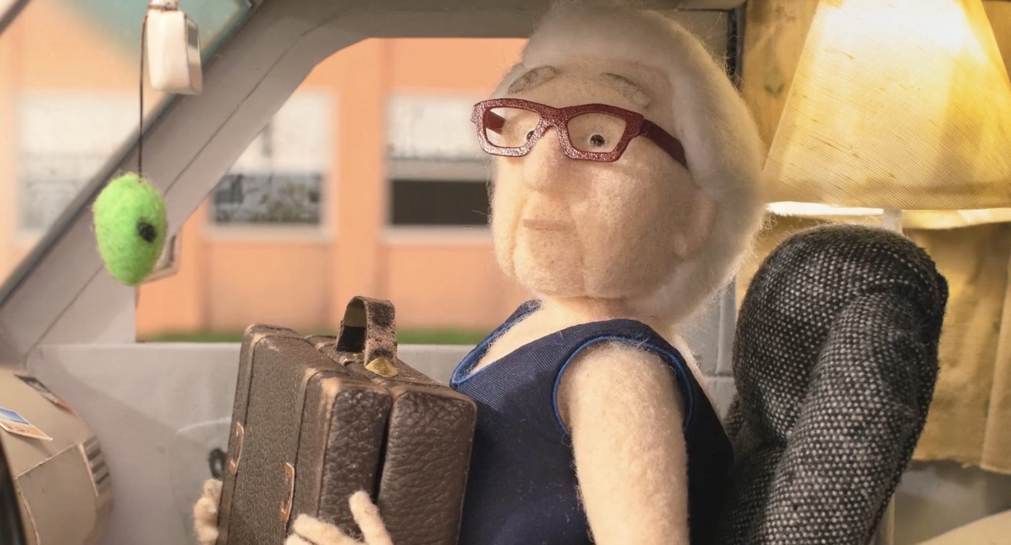 a still from the short animated film "Madeleine" by Raquel Sancinetti, featuring a stop-animation puppet of an elderly woman in a swimming suit, sitting in a car and holding a brown suitcase