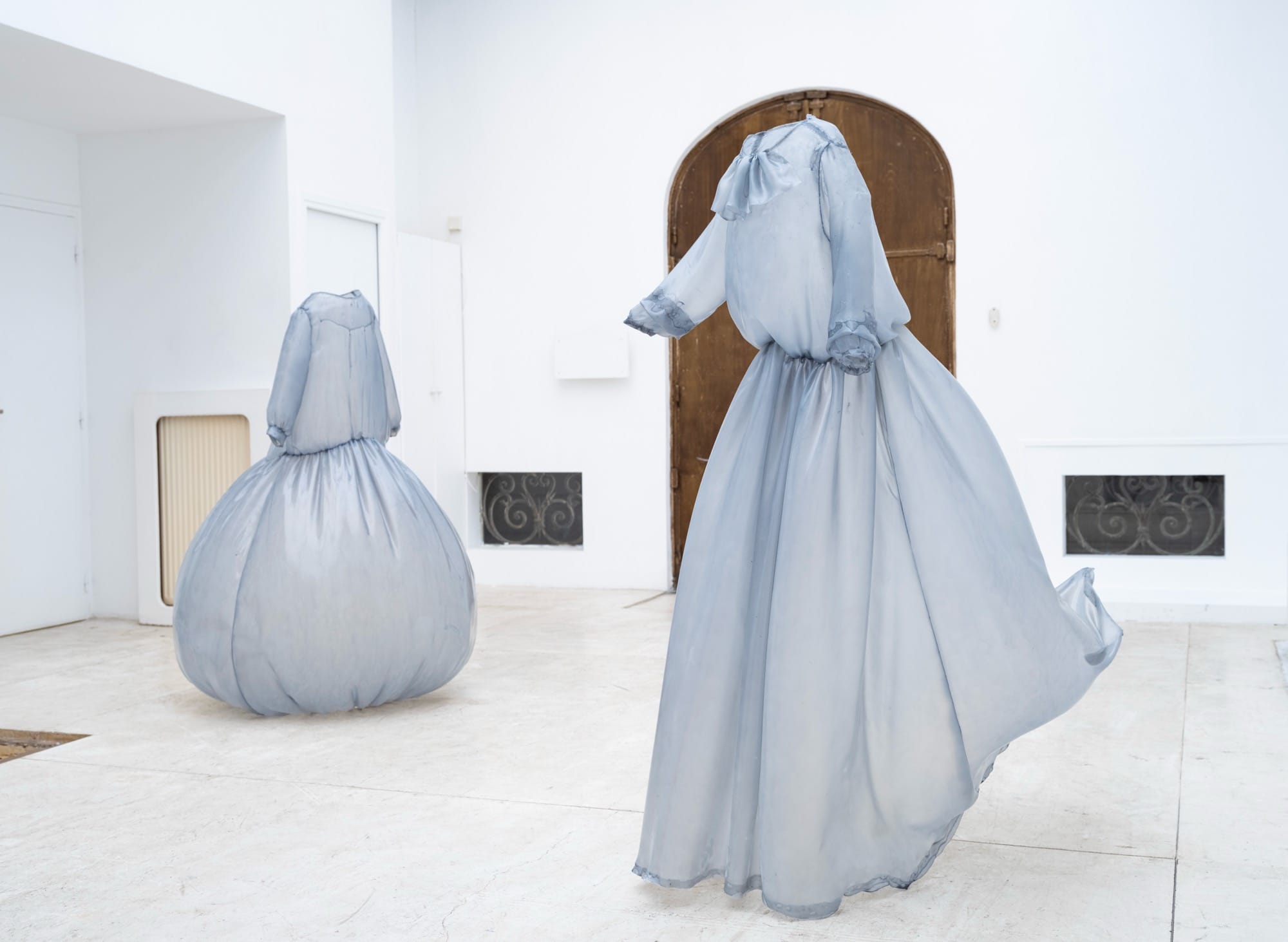 a collection of stiff pale blue fiberglass victorian style dresses by Brandon Morris that stand without wearers in the middle of a gallery