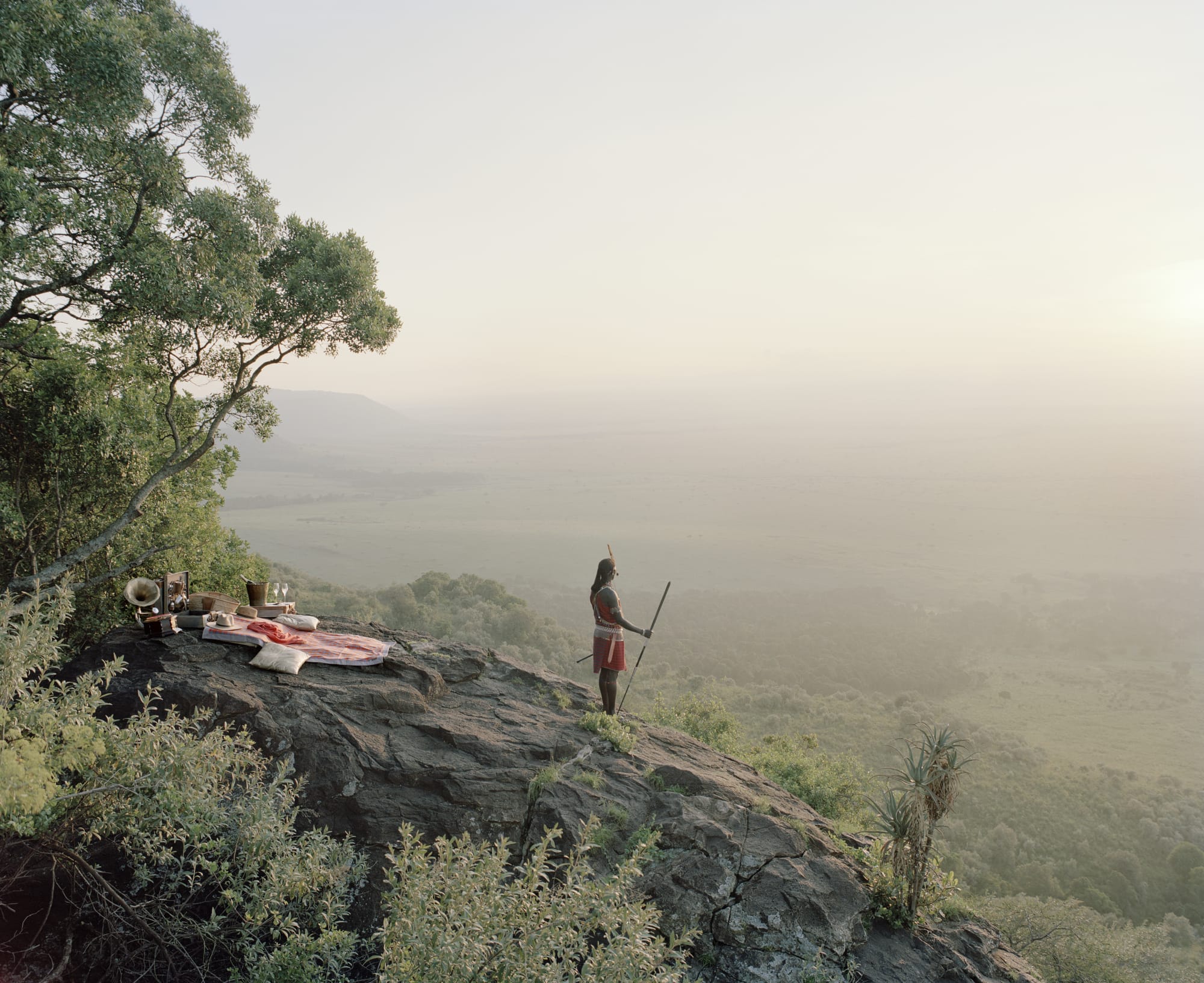 a Masai warrior stands on a hillside, looking out over a hazy landscape, with a pink picnic blanket on the rocks behind him