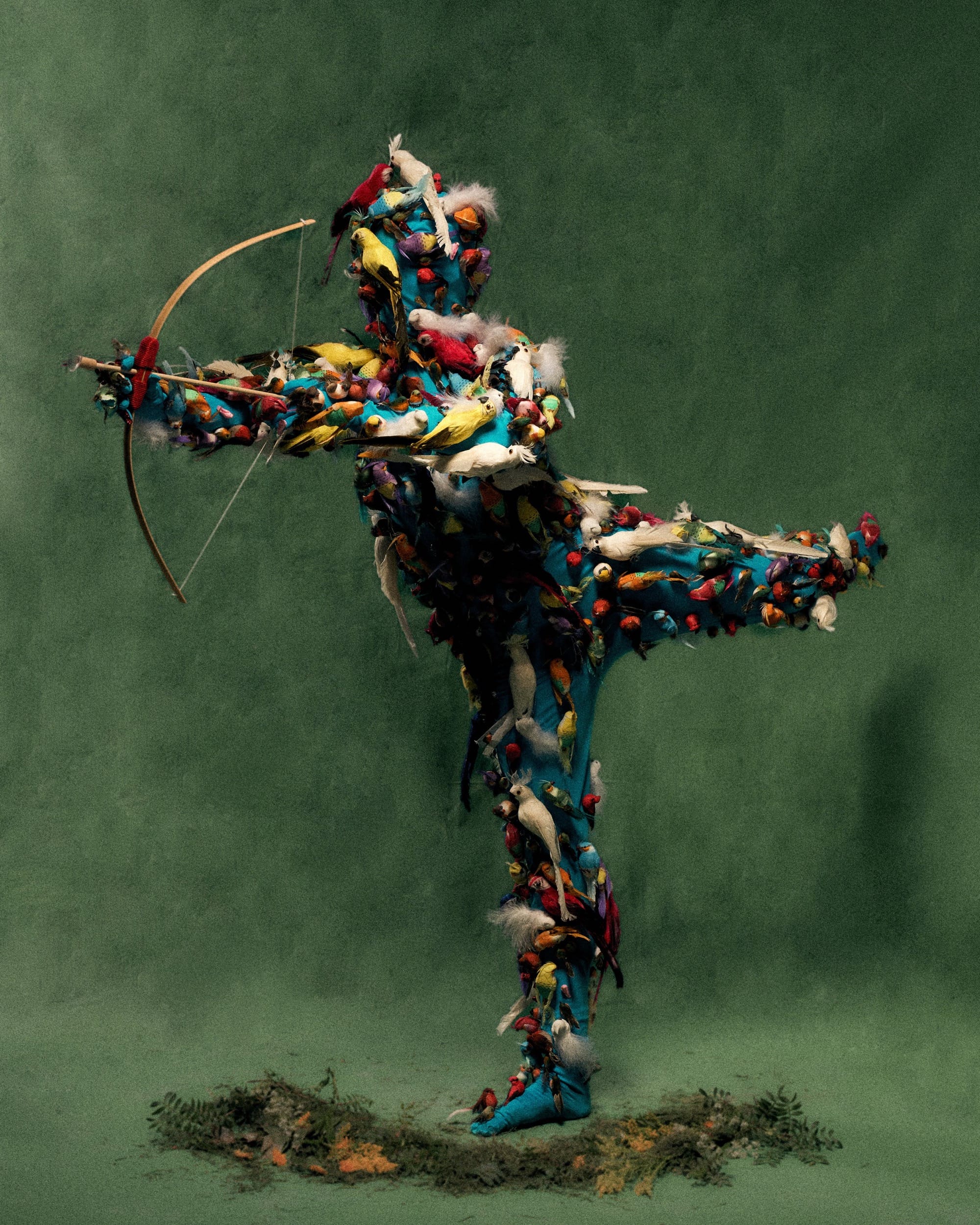 a costumed figure with an all-over outfit of faux birds, pointing a bow and arrow, posing against a green background